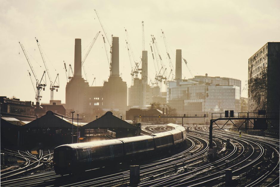 Photograph of the Battersea Power Station viewed from across the river, with four tall chimneys on the historic red brick building, surrounded by modern glass-fronted office buildings. In the foregrou
